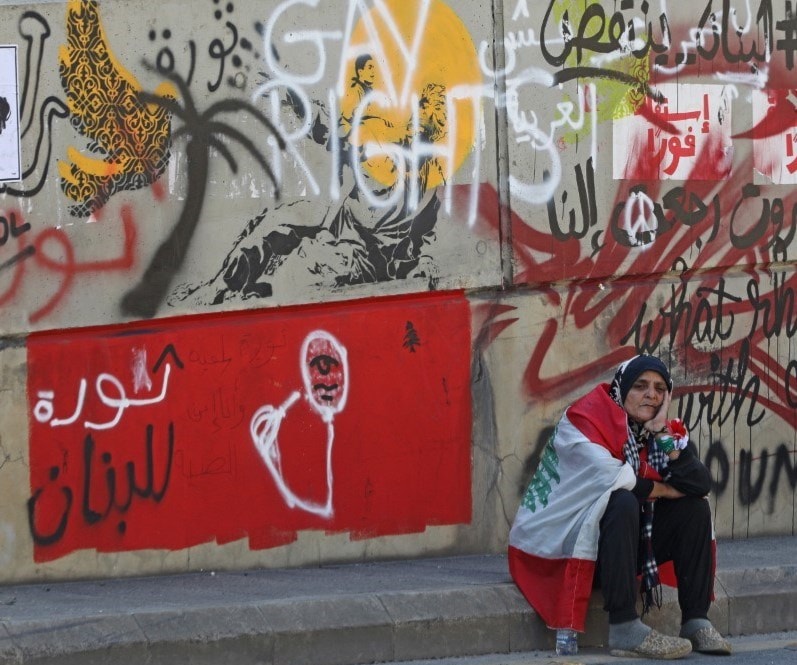متظاهرة لبنانية جالسة على رصيف أمام حائط ممتلىء برسومات وتعابير المتظاهرين في طرابلس (أ.ف.ب) متظاهرة لبنانية جالسة على رصيف أمام حائط ممتلىء برسومات وتعابير المتظاهرين في طرابلس (أ.ف.ب)
